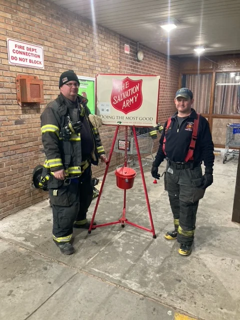 Two Belltown firefighters in turnout gear beside
                      a Salvation Army red-kettle stand outside a
                      storefront.