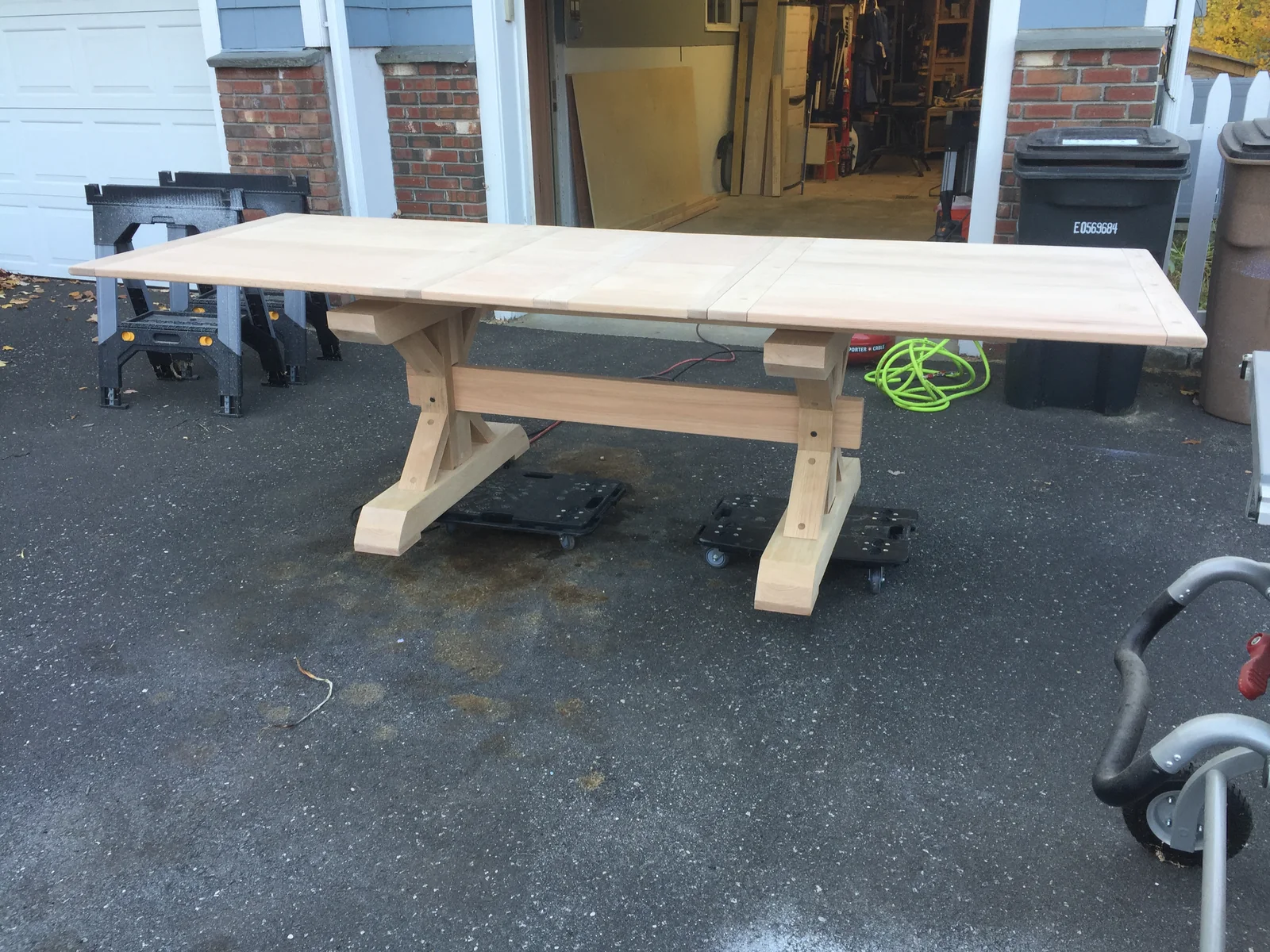 The assembled trestle dining table in bare,
                    unfinished wood, standing on dollies in the
                    driveway outside the garage workshop.