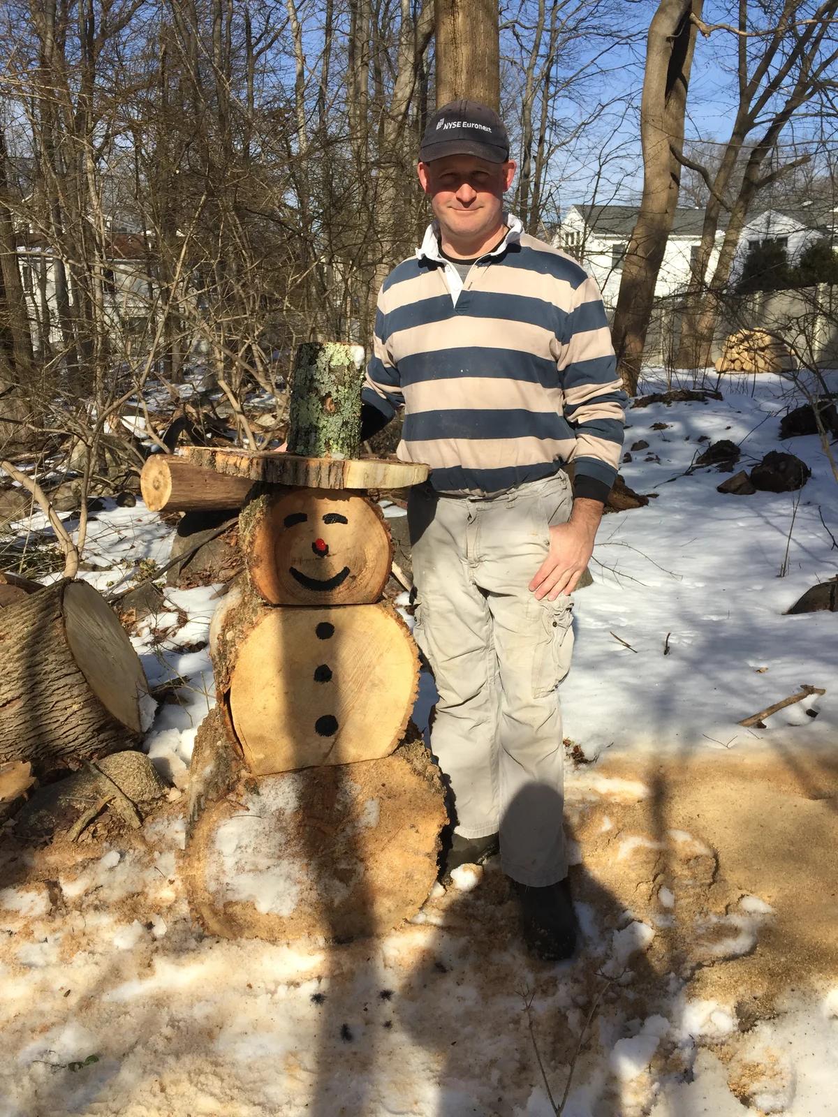 Scott standing in the snow beside a snowman-like
                    figure stacked from big log rounds, with a painted
                    smiley face, button dots, and a wood-slice hat.