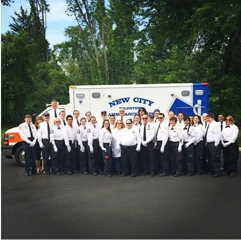New City Volunteer Ambulance Corps members in
                      uniform, posed as a group in front of an
                      ambulance.