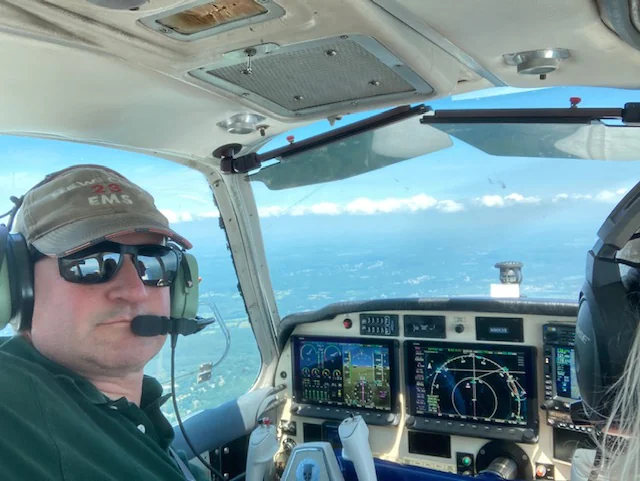 Scott at the controls of a light aircraft in cruise
                  flight, wearing a headset, with the instrument panel
                  and a hazy horizon of clouds visible through the
                  windscreen.